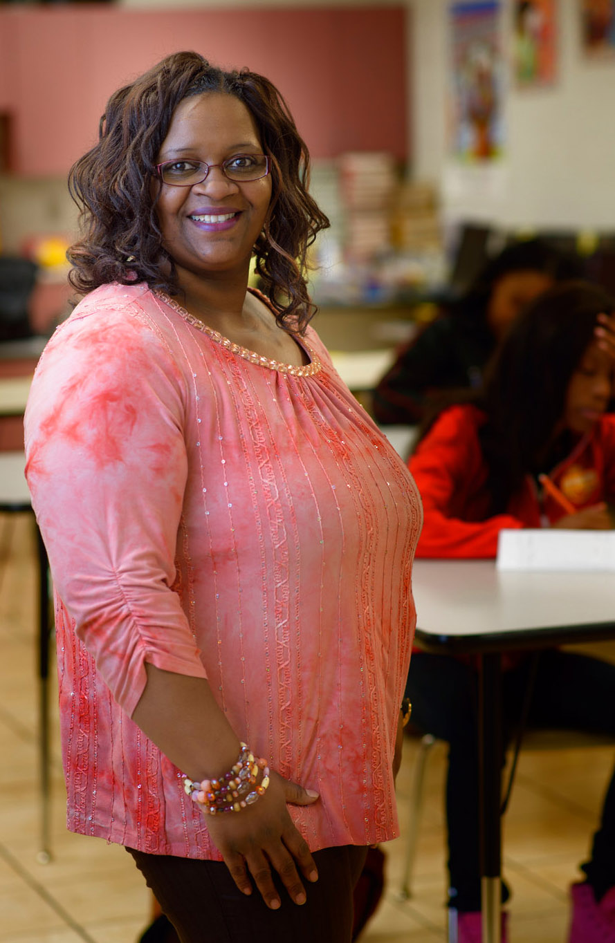 A woman with maroon-rimmed glasses and curly hair, wears a coral three-quarter sleeved blouse.