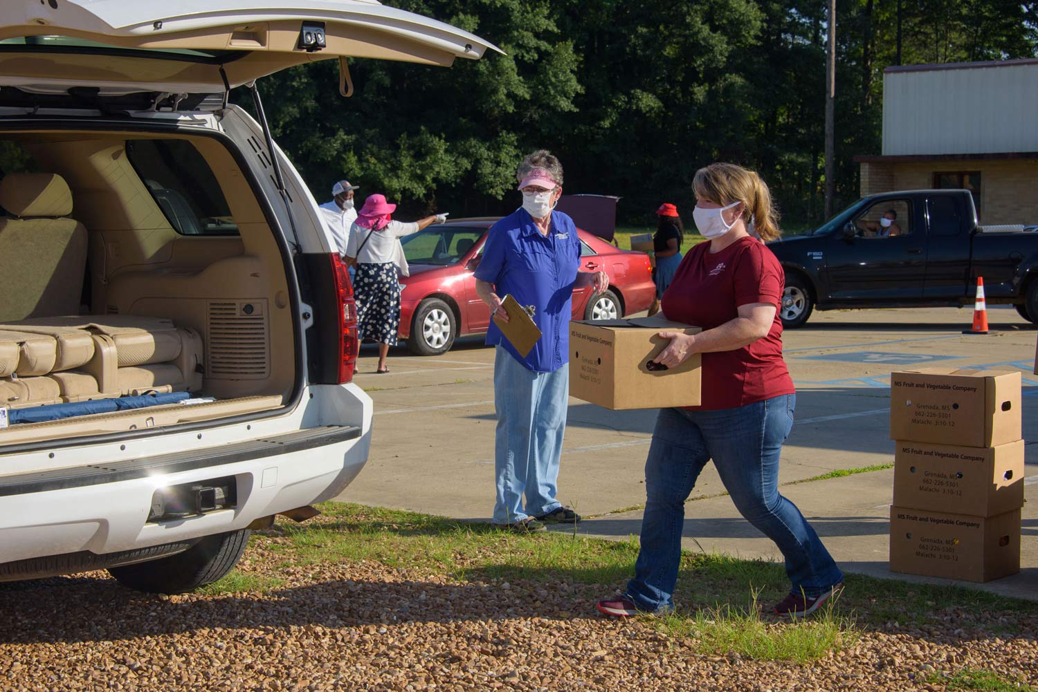 A woman wearing a mask carries a box to the open trunk of a white car.