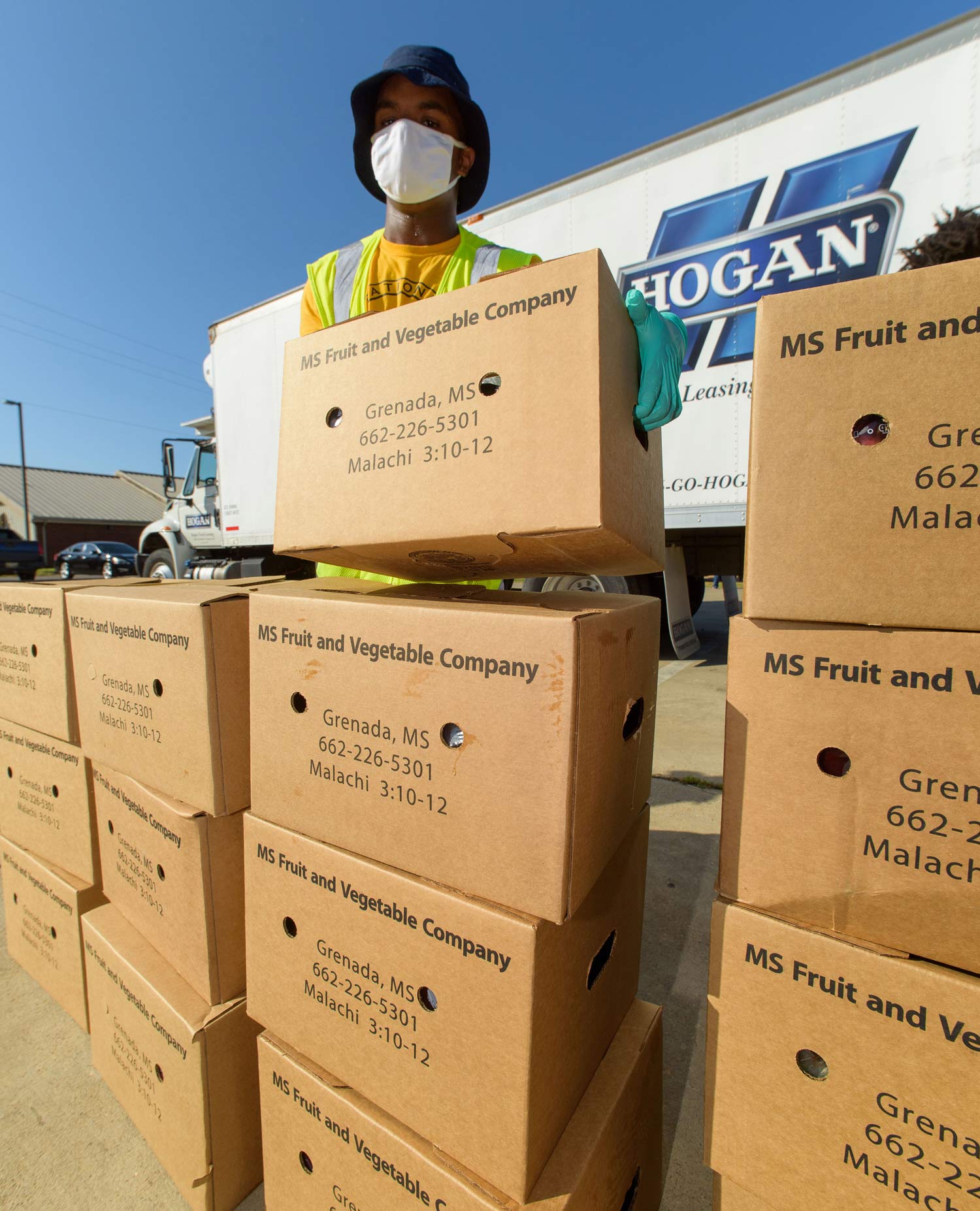 A man wearing a mask stands behind a tall stack of cardboard boxes labeled “MS Fruit and Vegetable Company.”