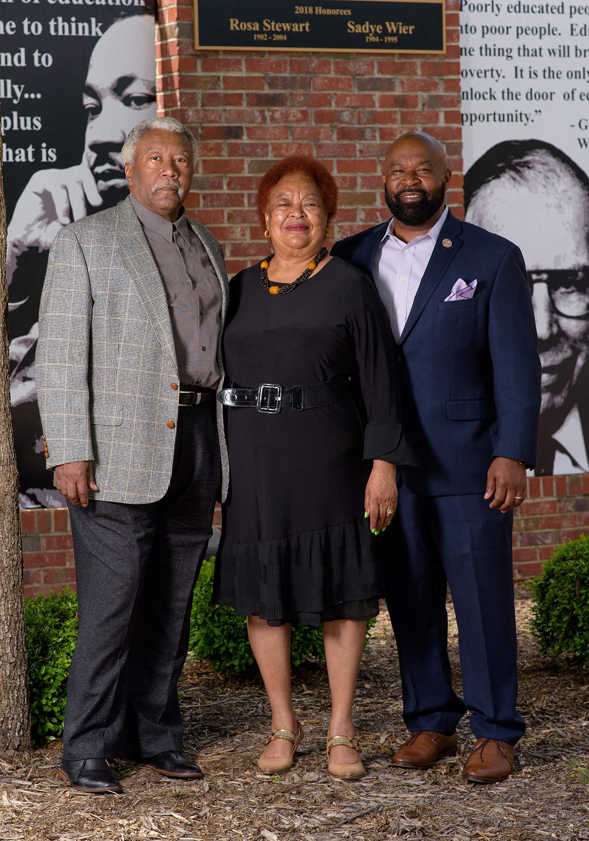 An older man and woman with a younger man stand smiling in front of a sign honoring Sadye Weir.