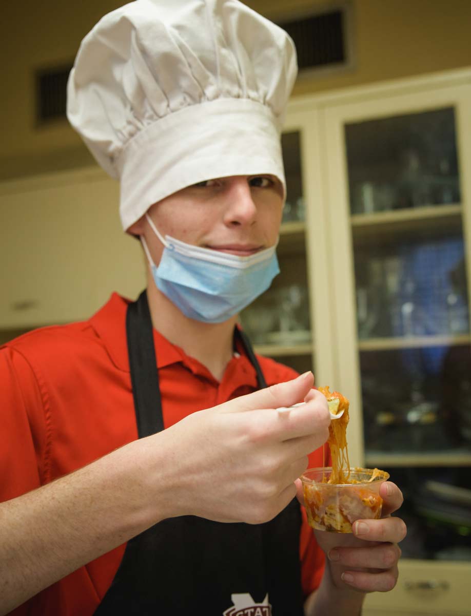 A young man wearing a chef’s hat tasting a cup of noodles.