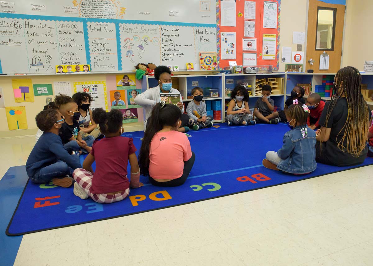 A young woman reads to a group of small children sitting on a blue rug in a classroom.