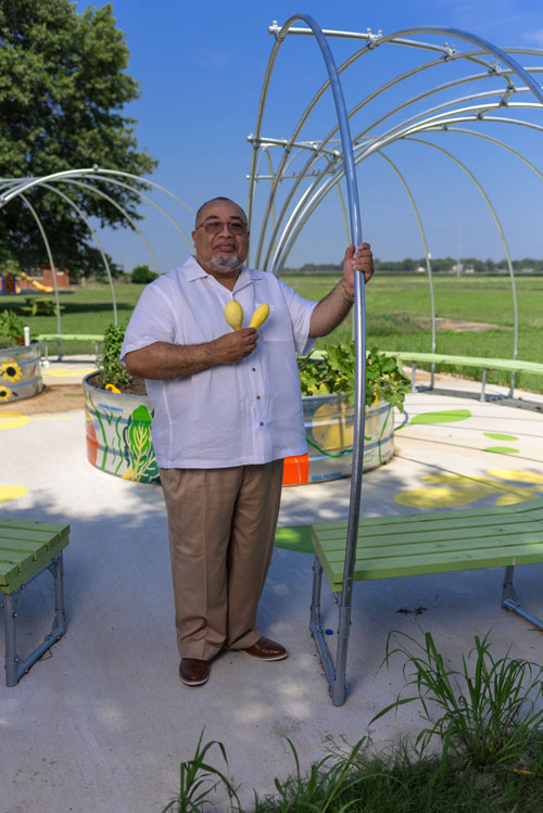 A man holding two small yellow squash stands outside in a garden.