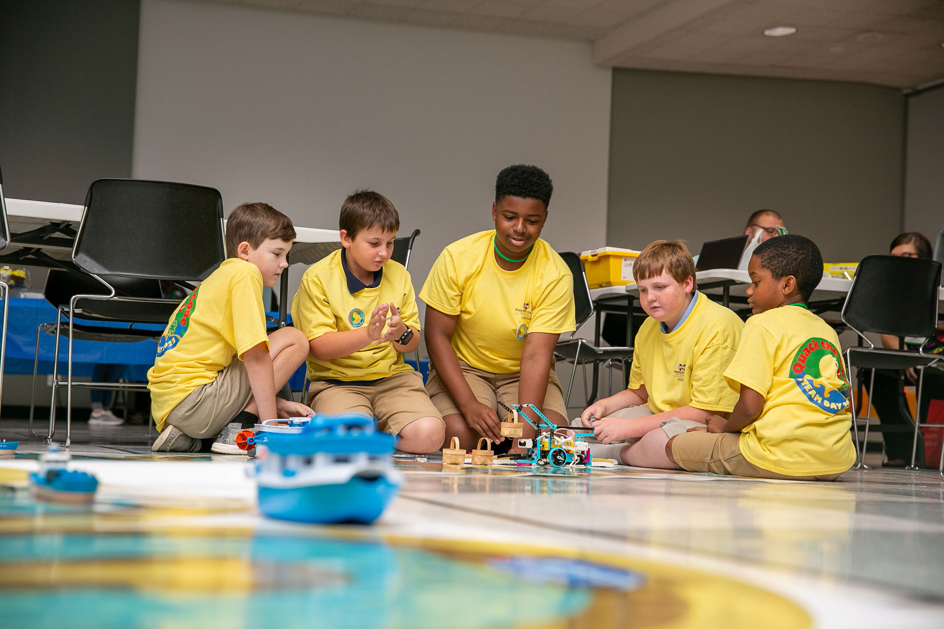 Five boys wearing yellow T-shirts surrounding a robot picking up small baskets.