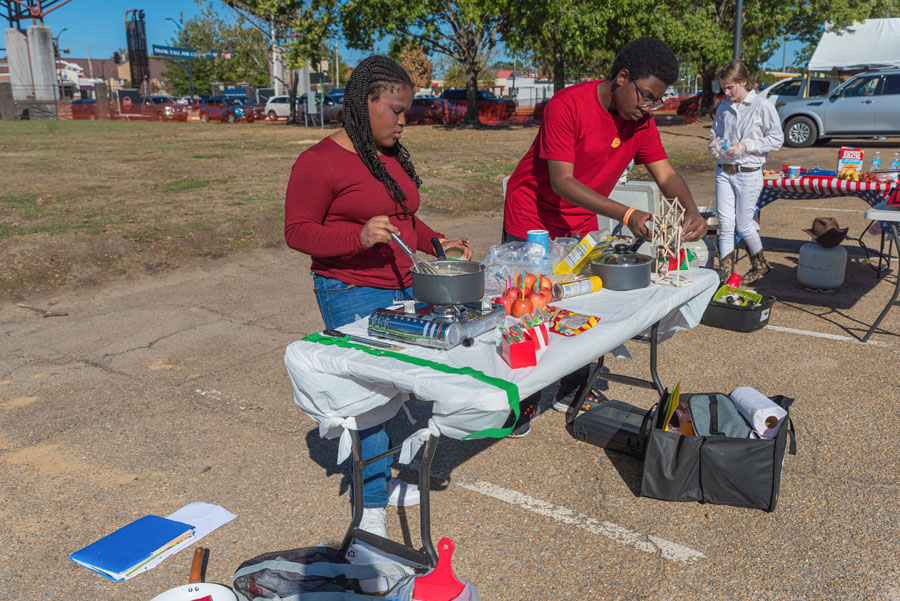 A girl and a boy, both wearing red shirts, stir cooking pots on a table outside.