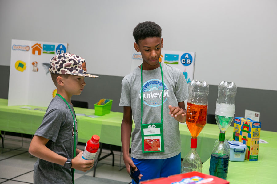 A white boy and black boy look at two upside down liter bottles, one filled with orange liquid and the other with clear.