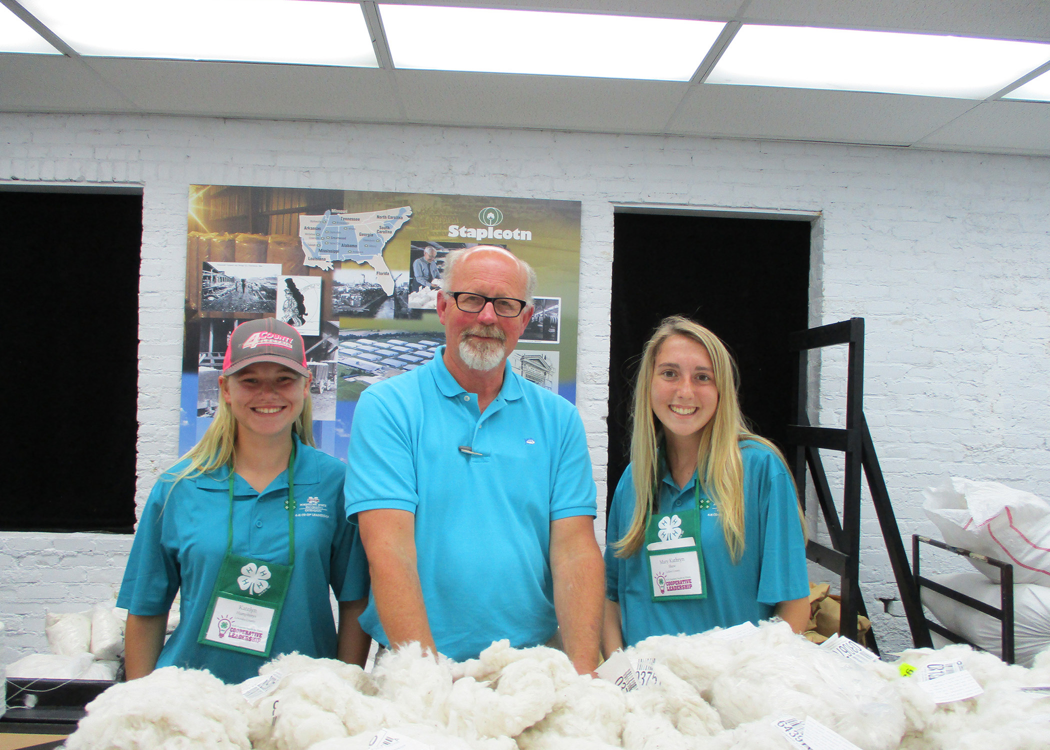 Two blonde haired girls and a middle age man all wearing blue shirts pose next to a table with cotton on it.