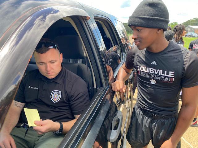An officer holds a ticket while sitting in a vehicle. A young person is peering through the officer’s rolled-down window.