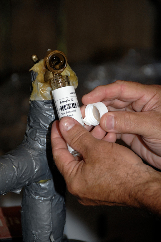 A person collects a water sample in a small container with a barcode on its label.