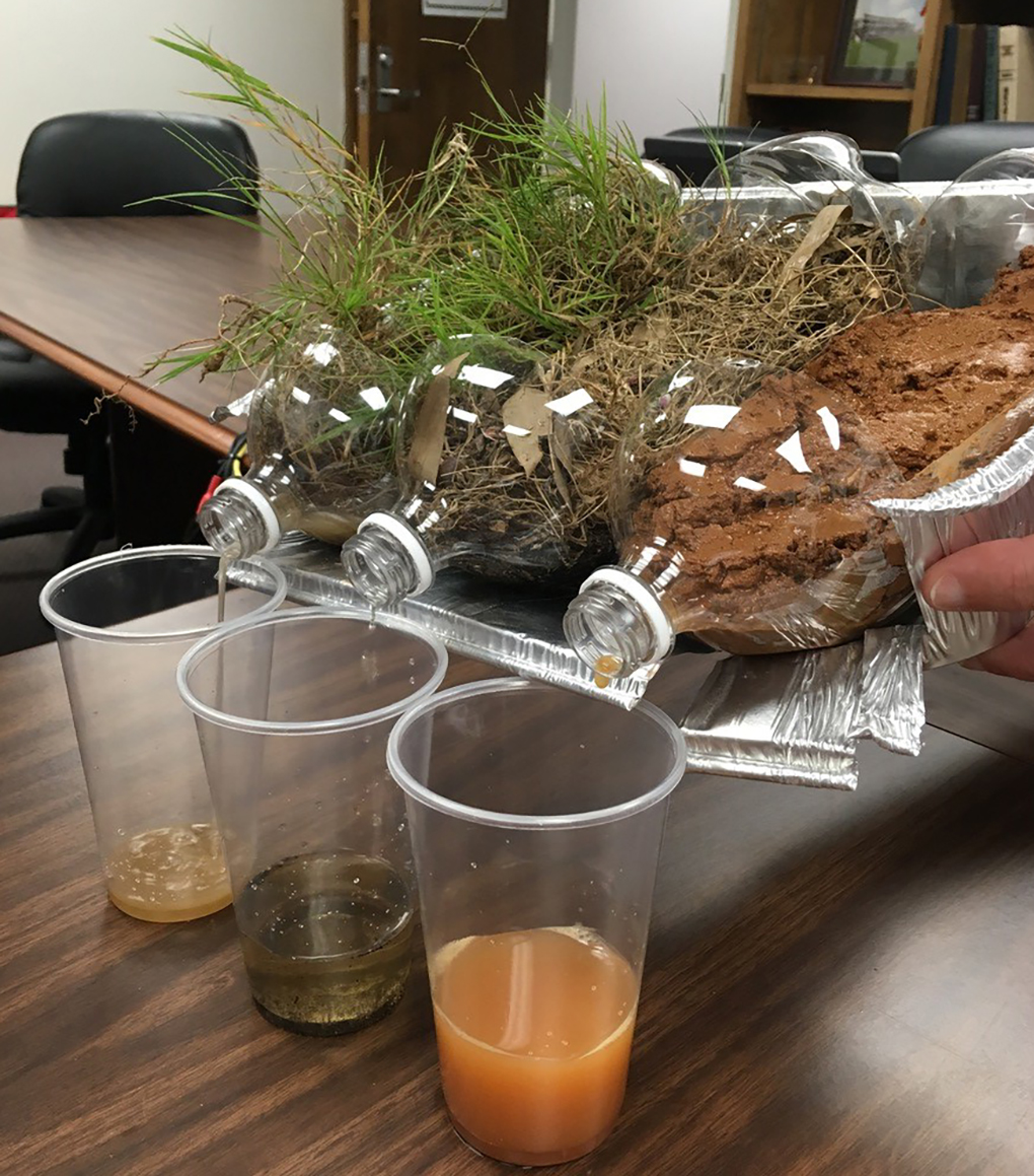 Three 2 liter bottles leaking water into three plastic cups, simulating rainfall.