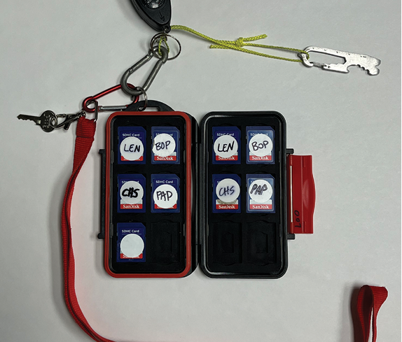 An SD case with one unlabeled SD card on the left side and four labeled SD cards on the right. The case, keys to the cable lock, and multi-tool are attached to a blue lanyard with an American flag image on it.