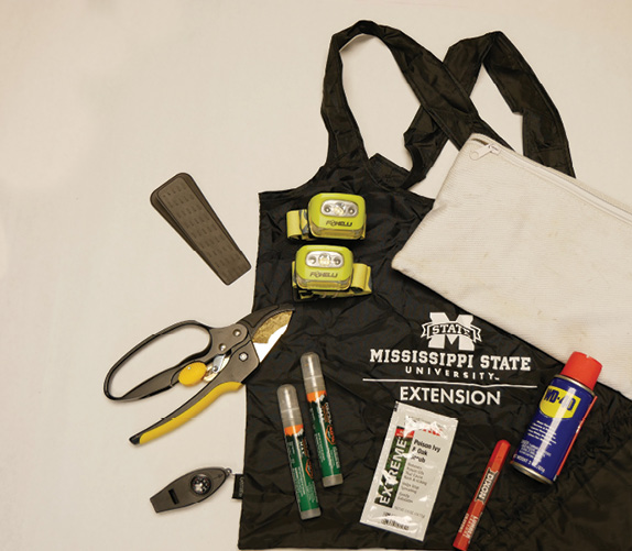 A layout of items on top of a black Mississippi State University Extension logo field bag: a doorstop, two small head lamps, shears, a compass and security whistle, two insect repellent pens, a packet of poison ivy gel, a red lumber crayon, a can of WD-40, and a canvas kit bag.