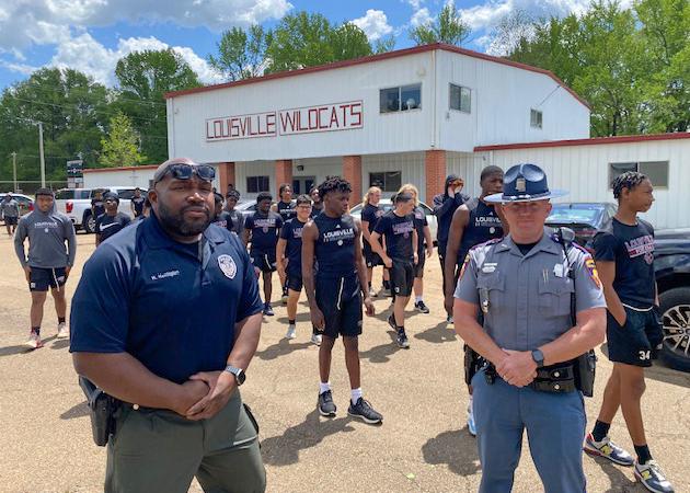 Two officers stand in front of a group of young men spaced apart for workouts in a parking lot.