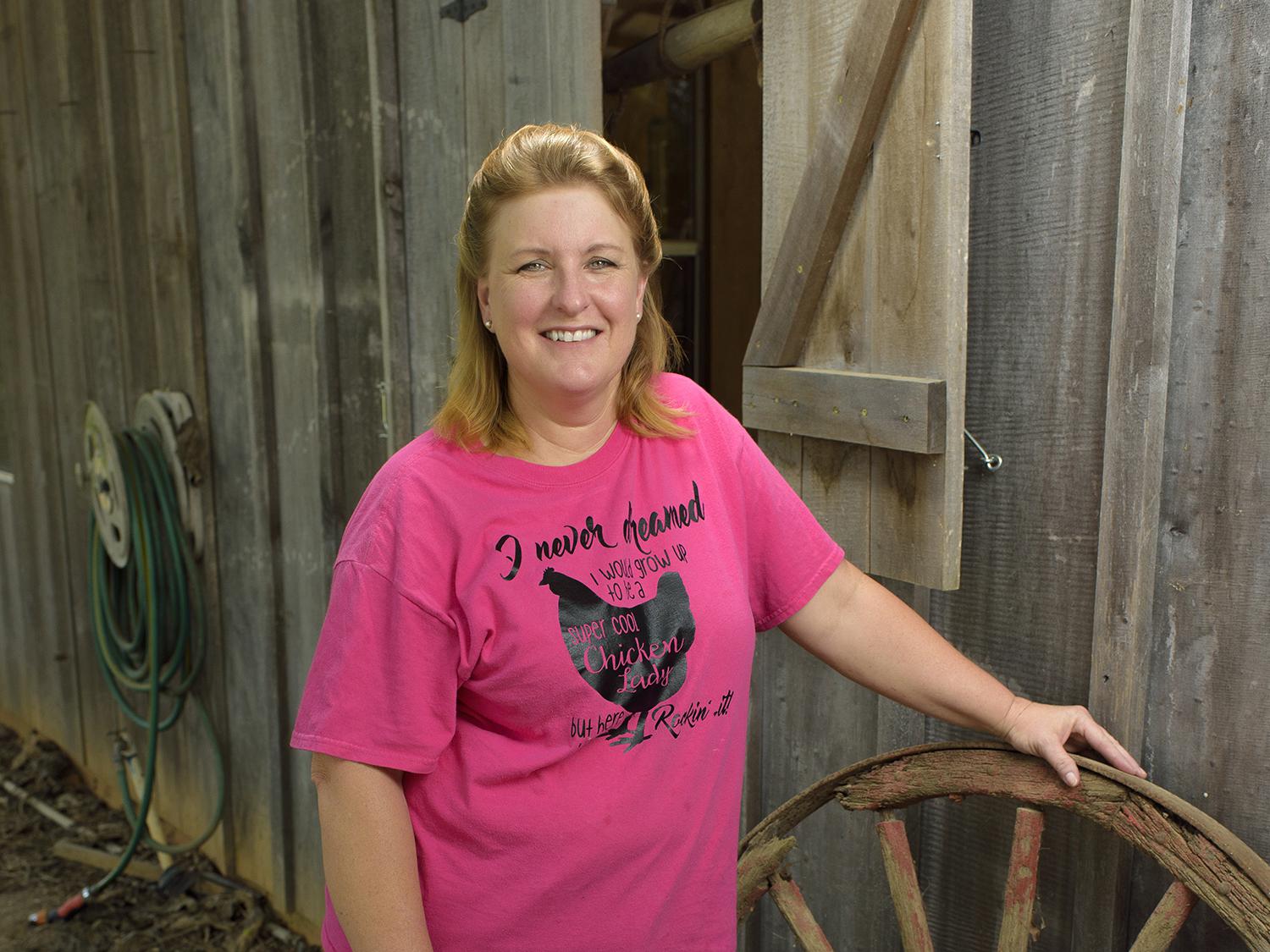 Dressed in a pink T-shirt and blue jeans, broiler grower Teresa Dyess stands next to two wagon wheels in front of a barn on her family farm.