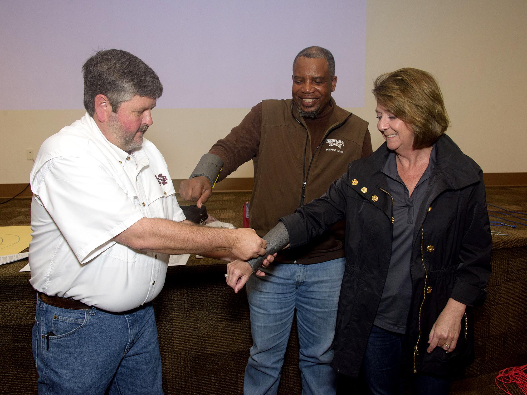 A man mimics removing a fishing hook from a woman’s arm during a safety demonstration as another man looks on.