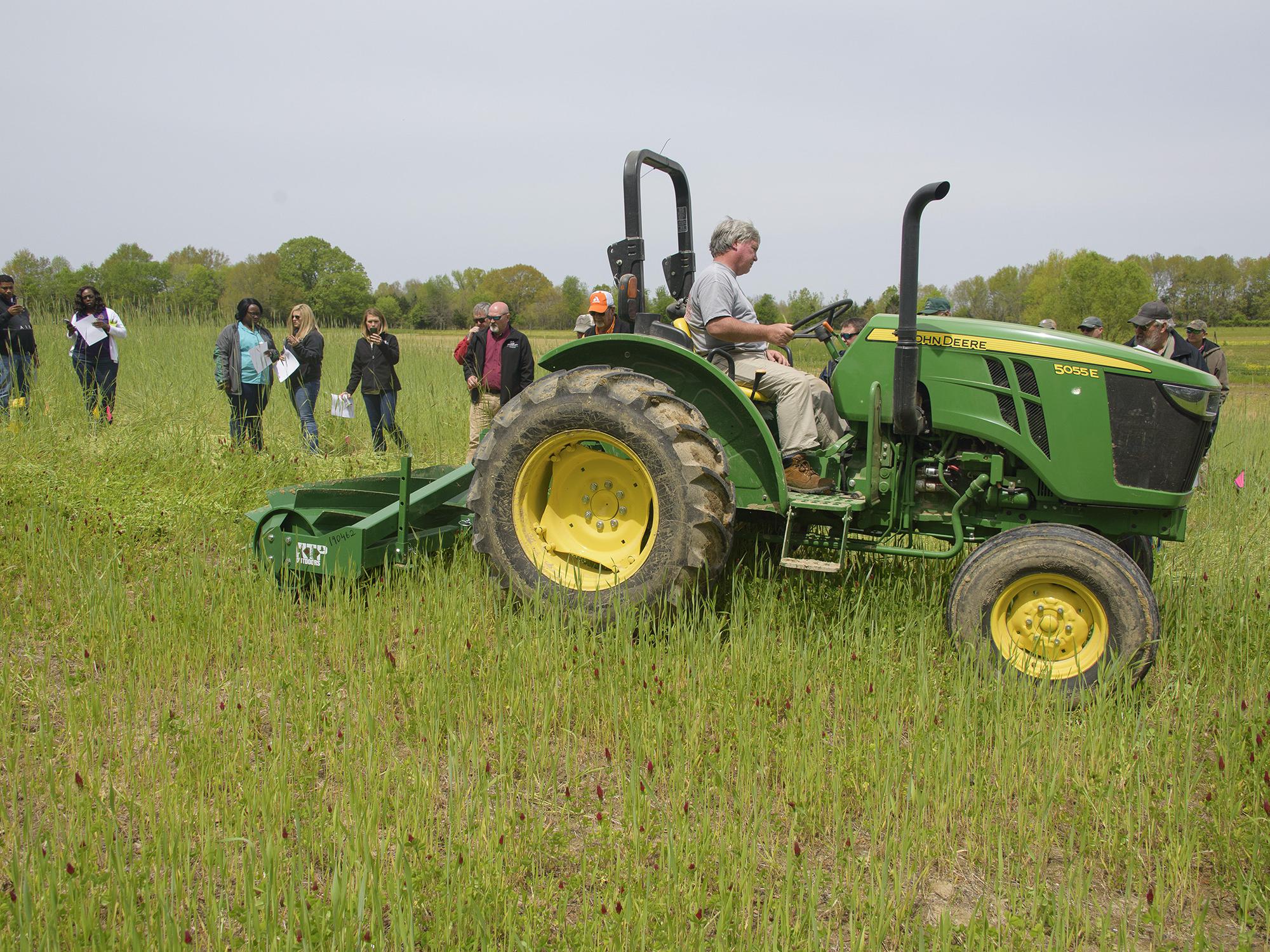 Man drives tractor pulling a large roller over a grass field while spectators watch.