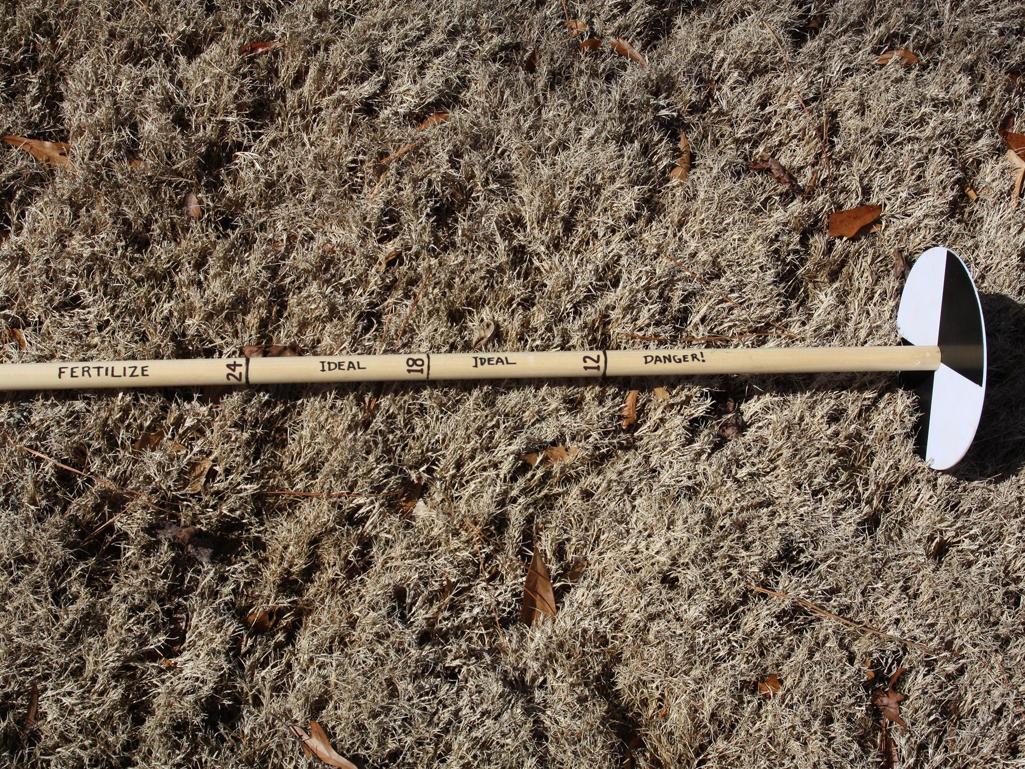 A wooden pole with hash marks at 12, 18 and 24 inches from a round disk with alternating black and white quadrants rests on brown grass. Danger is written below the 12-inch hash, ideal is written in the middle section, and fertilize is written above the 24-inch hash.