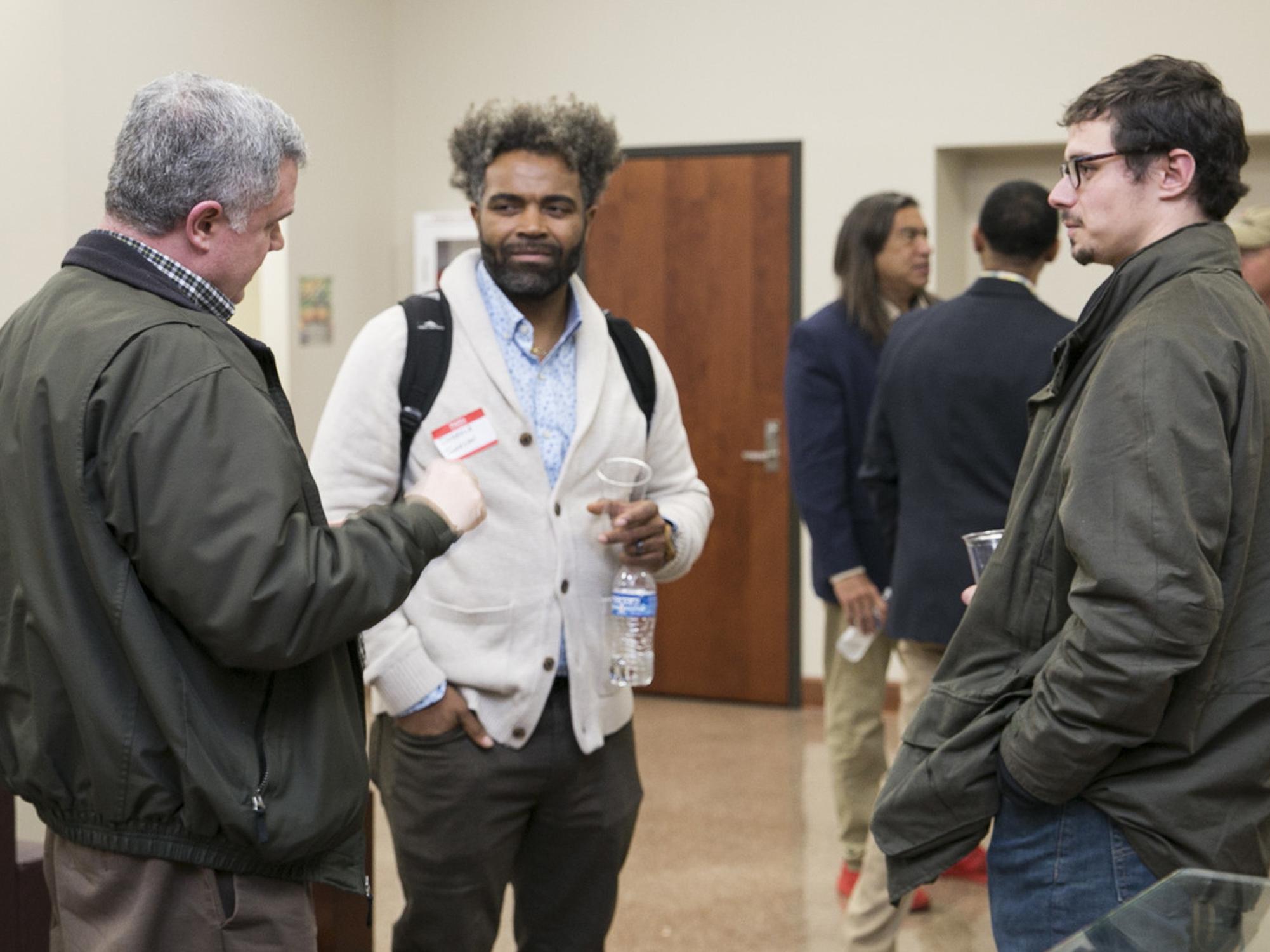 Three men talk together in a group, with several others clustered in the background.