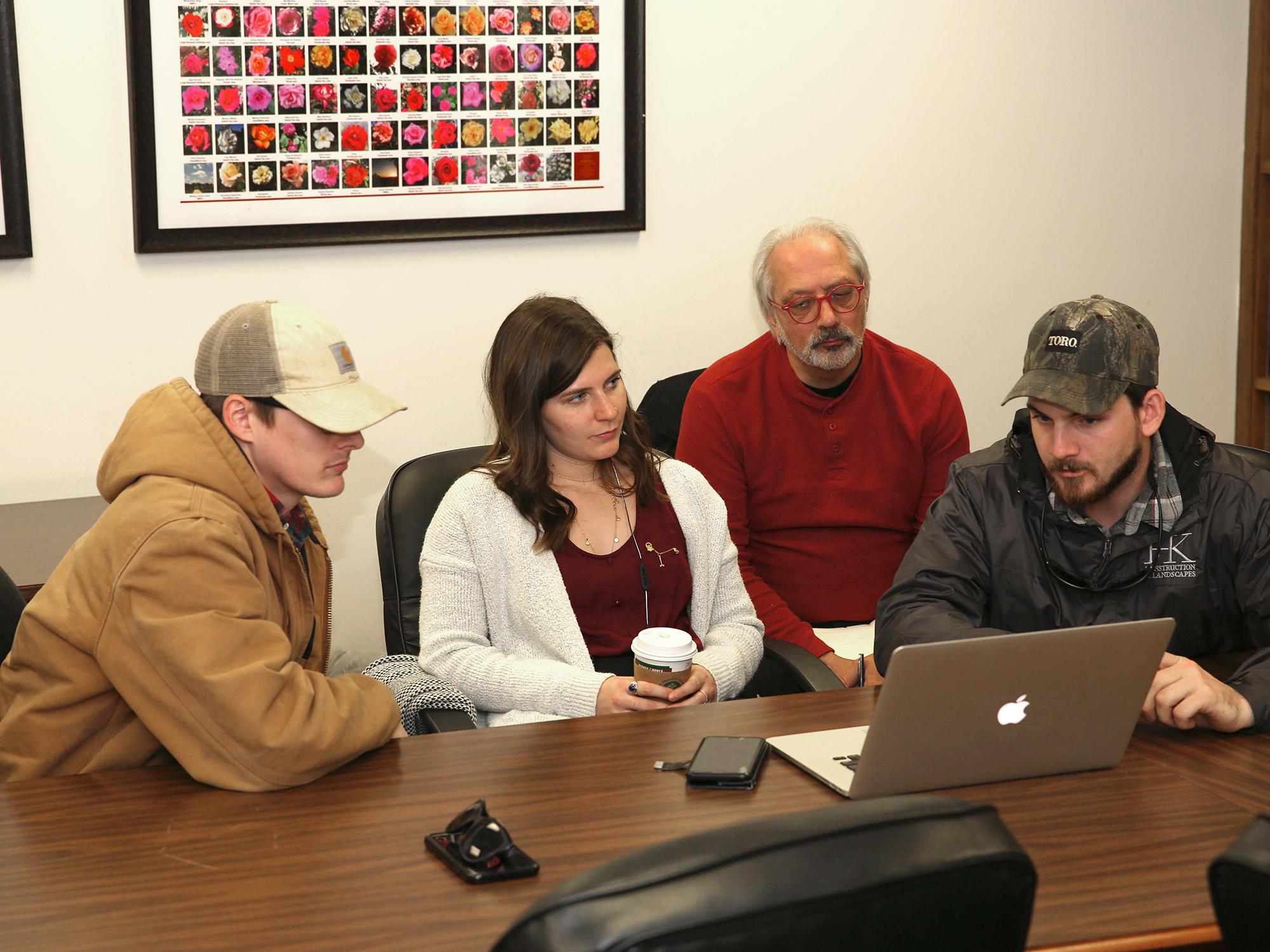 A group of three students gather around a computer while a man looks on from behind.