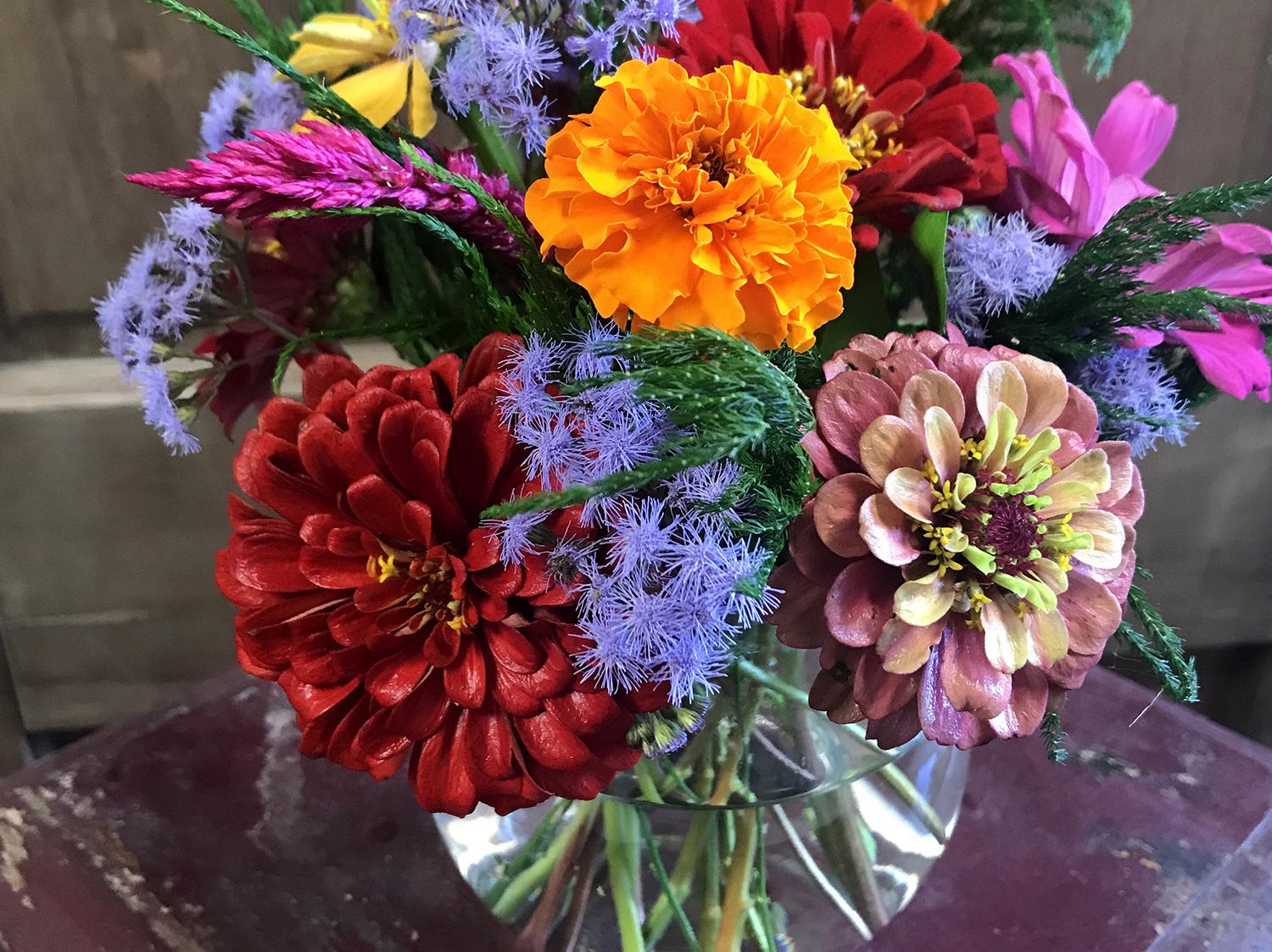 Orange, red, pink, purple, and yellow flowers are arranged in a glass bowl that sits atop a small, red table.