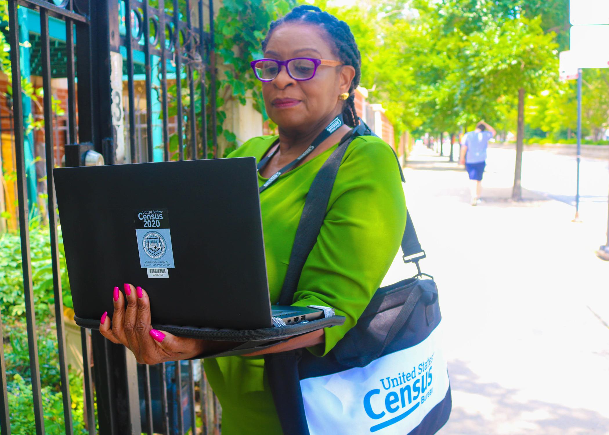 Woman in a green dress with a bag around her shoulder stands in front of a gate holding a laptop computer.