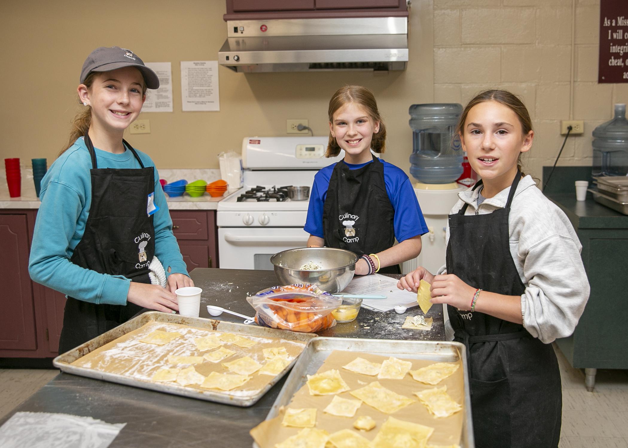 Three girls work with dough sheets at a table.