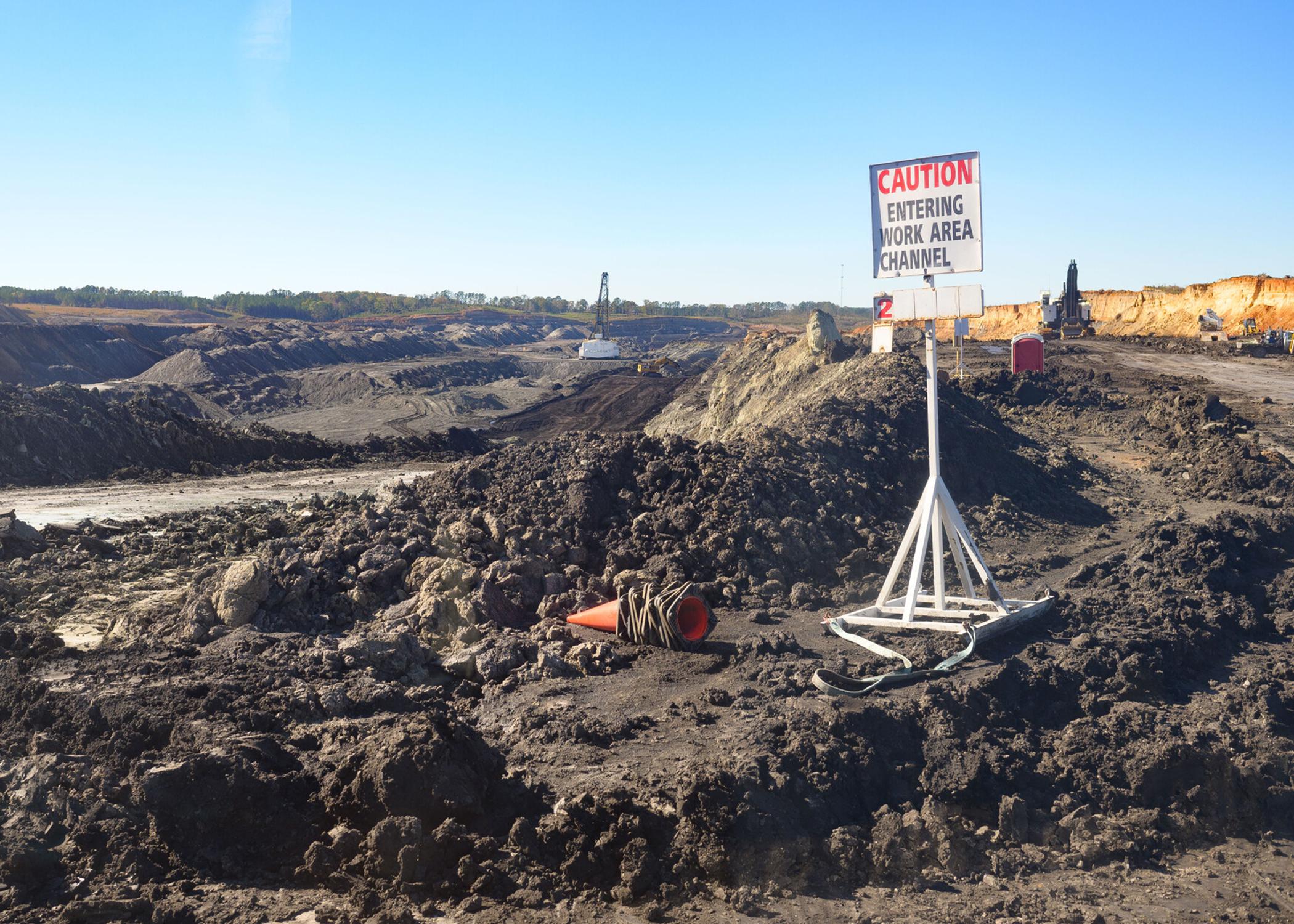 Wide shot of a surface coal mine featuring a layered landscape of exposed rock and soil, heavy machinery, and a sign that reads “caution, entering work area channel.