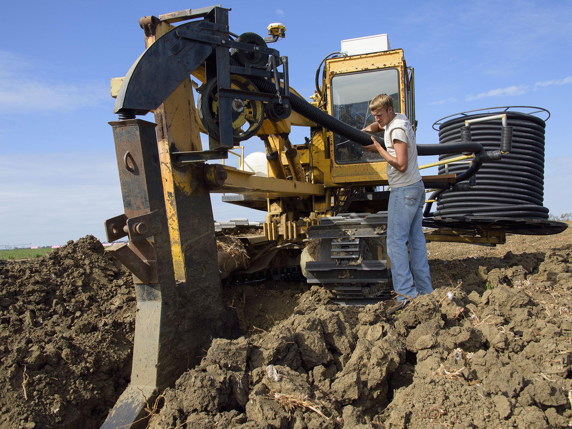 Dylan Yost feeds plastic tubing into equipment that buries drain tile in a deep furrow in fields. This Noxubee County field was being tiled Nov. 8, 2016. (Photo by MSU Extension Service/Kevin Hudson)