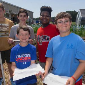 Two boys outside, holding paper towels with seeds, and three boys in behind them, one holding a shovel and another with a paper towel, all smiling.