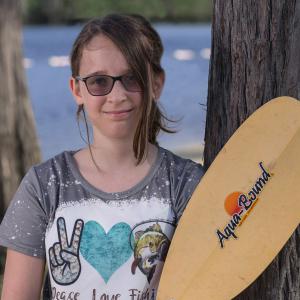 A teen girl smiling, wearing sunglasses and holding a rowing oar.