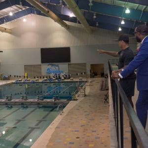 Two men look over a swimming pool, and one extends his hand toward the water.