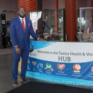 A man wearing a suit stands beside a desk with a banner listing “Welcome to the Tunica Health & Wellness Hub.”