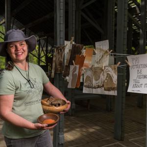 A smiling woman, holding a bowl of rocks in one hand and a bowl of sandy colored dirt in the other, standing in from of paintings hung on a line to dry.