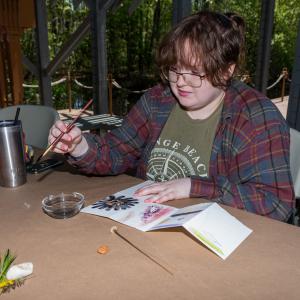 A woman holding a paint brush and looking at a canvas with flowers and abstract art on it.