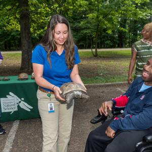 A person seated in a wheelchair smiles at a turtle being held by another person.