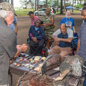 A group of people watch a man at a table with mock turkeys and real turkey feathers.