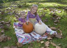 Two young girls sit on a colorful quilt among leaves in the grass as they play with a white and an orange pumpkin.