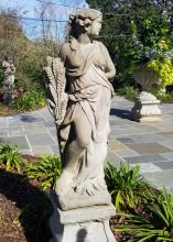 Stone statue of a female holding harvested wheat stands in a landscape bed.