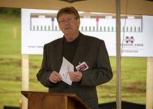 A man in a grey blazer stands behind a podium and speaks.