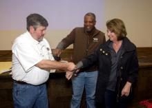 A man mimics removing a fishing hook from a woman’s arm during a safety demonstration as another man looks on.