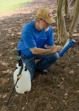 A man kneels on the ground beside a pump sprayer to read the label on the back of a pesticide jar.