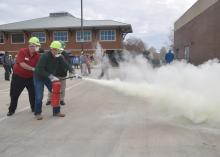 Two men wearing hard hats and masks activate a fire extinguisher as MyPI training participants watch. 