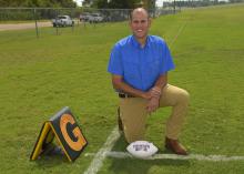 A man in a blue shirt kneels on a chalk-lined football field beside a goal line marker. A white Mississippi State football rests on the ground beside his knee.