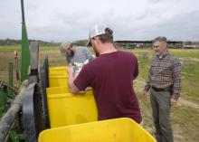 Two young men pour seed into bright yellow bins while a man watches.