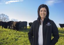 A woman looks into the camera as a herd of black cows feed in a group behind her.