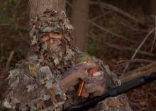 Only the eyes of a turkey hunter wearing full camouflage is visible. He is holding a wooden turkey caller.