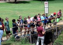 More than a dozen young people listen to a teacher while they stand on a cement bridge overlooking a large ditch.