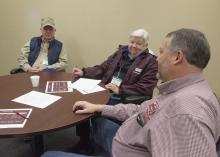 Man and woman sit at a round table with papers on it while they look toward another man beside them at the table.