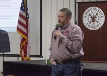 Man speaks into a handheld microphone. Background includes the Mississippi State University seal and an American flag.