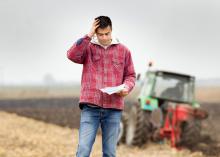 A farmer stands with a tractor in the background looking at a document and holding a hand to his head in worry.